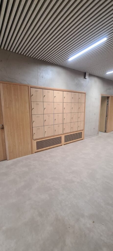 Mur de casiers vestiaires en stratifié compact HPL finition chêne naturel, cases numérotées de 097 à 119, équipés de serrures à clé à bec discret, intégrés sur mesure dans un couloir avec plafond à lames bois et sol béton ciré