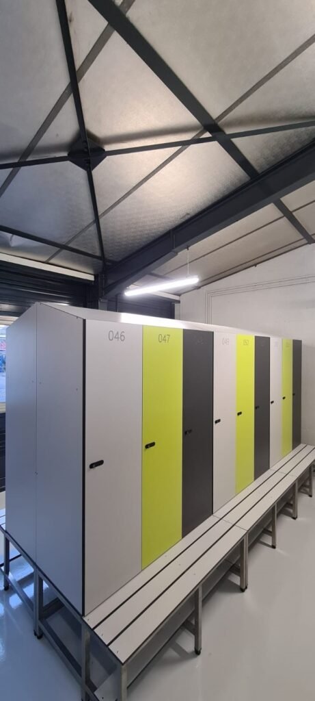 Rangée de casiers vestiaires pleine hauteur en stratifié compact HPL bicolore gris clair et vert anis, numérotés de 046 à 051, équipés de serrures à clé, accompagnés de bancs vestiaires en HPL sur pieds métalliques, dans un vestiaire sportif sous charpente métallique